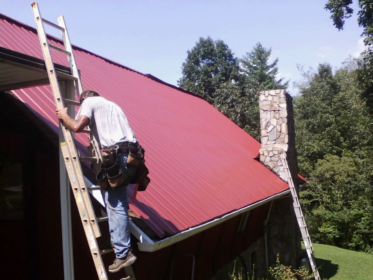 Roofer on a ladder beside a red metal roof and stone chimney — Soffit & Fascia Repair by Roofing Brothers in Americus